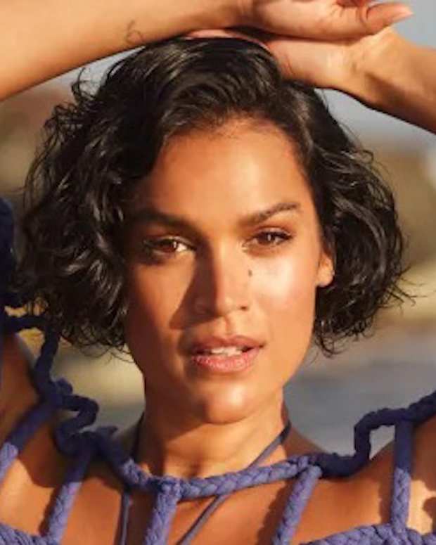 Jennifer Atilémile poses in a blue swimsuit and braided cover-up with her arms above her head,